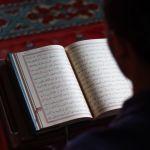 Mushaf Al-Quran terbuka di atas rehal dengan seseorang membaca di dalam masjid